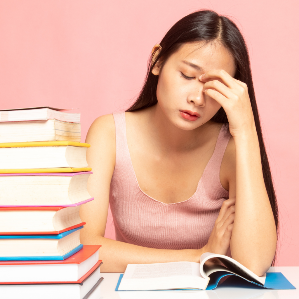 Frau überfordert, müde, antriebslos und mental ausgelaugt durch zu hohen Zuckerkonsum. Sitzt vor einem Stapel Bücher.