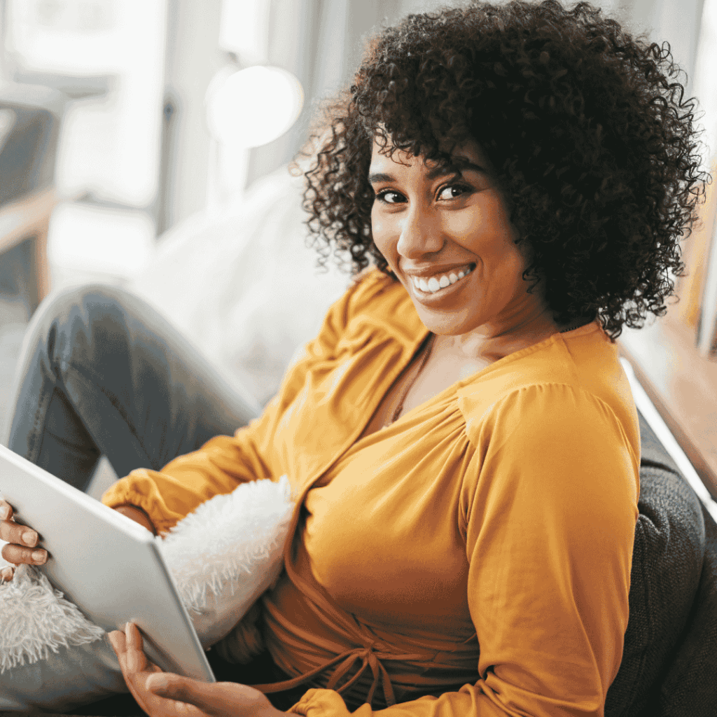 Lächelnde Frau in gelber Bluse sitzt entspannt mit Tablet auf dem Sofa – Symbol für Selbstfürsorge, achtsames Zeitnehmen und bewusste Pausen im hektischen Alltag.