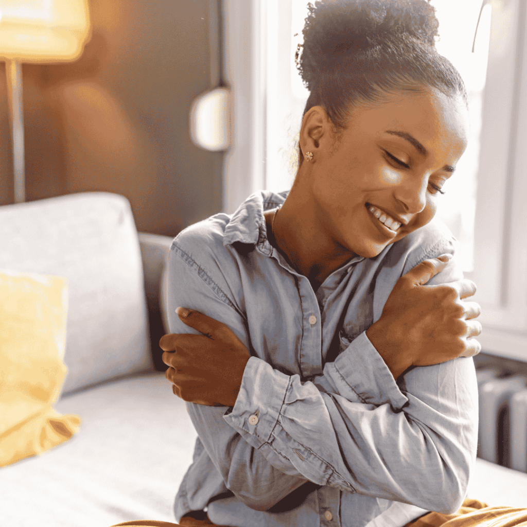 Frau sitzt lächelnd auf dem Sofa und umarmt sich selbst – Ausdruck von Selbstliebe, innerem Frieden und achtsamer Verbindung mit sich selbst im Alltag.