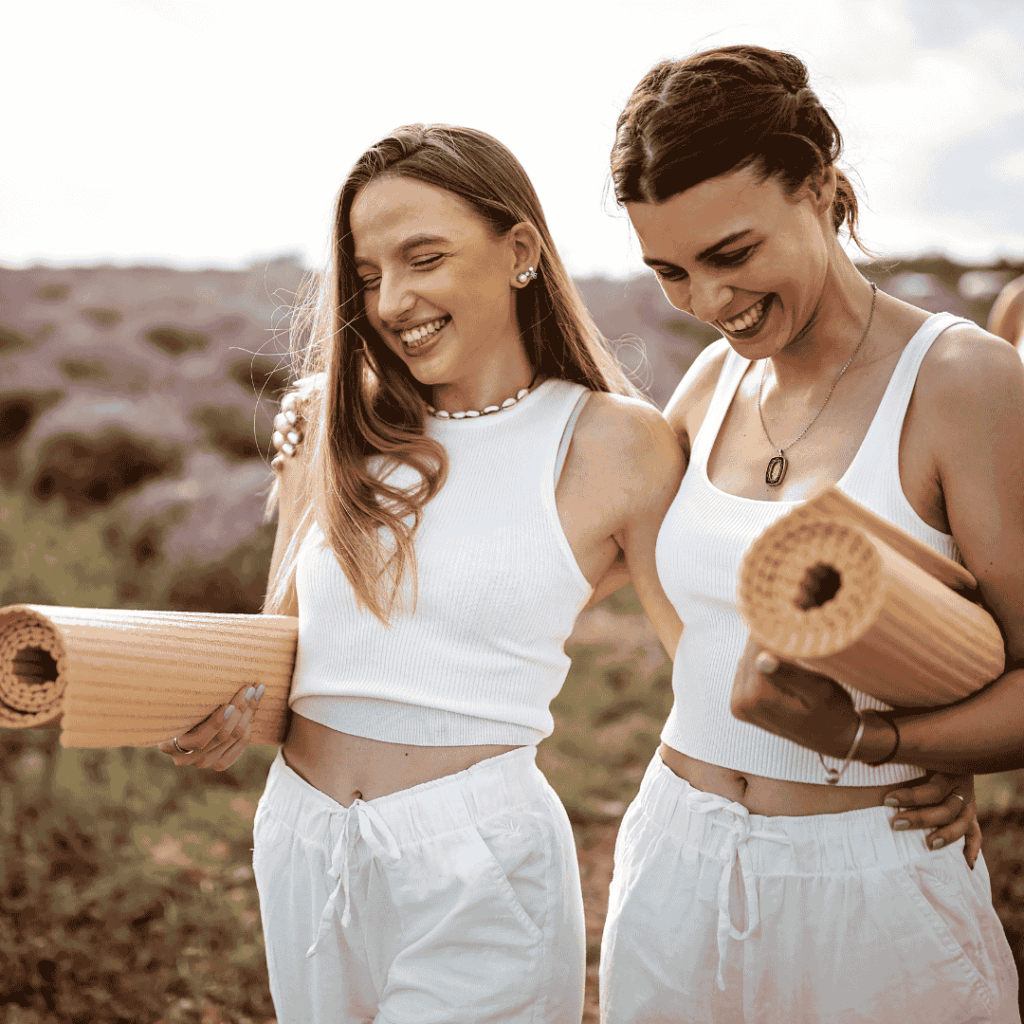 Zwei junge Frauen in weißer Yoga-Kleidung lachen gemeinsam in der Natur, mit Yogamatten im Arm – ein Moment voller Selbstliebe, Leichtigkeit und Anti-Stress-Energie.