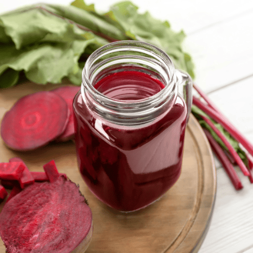 Ein hormonfreundlicher Anti-PMS Saft mit Roter Bete in einem Glas mit Henkel auf einem Holzbrett, umgeben von frischer Roter Bete und Blättern – ideal als zyklusfreundlicher Saft zur Linderung von PMS.