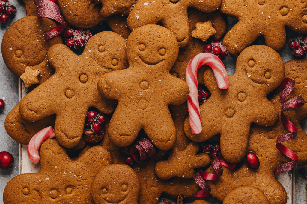 Nahaufnahme eines Blechs mit frisch gebackenen veganen Lebkuchen Plätzchen ohne Zucker, in Form von Lebkuchenmännchen. Mit Kokosmehl wie Schnee bestäubt, dekoriert mit roten Beeren, Zuckerstange und weihnachtlichen Gewürzen. Gemütliche Boho-Weihnachtsstimmung, warme Farben.