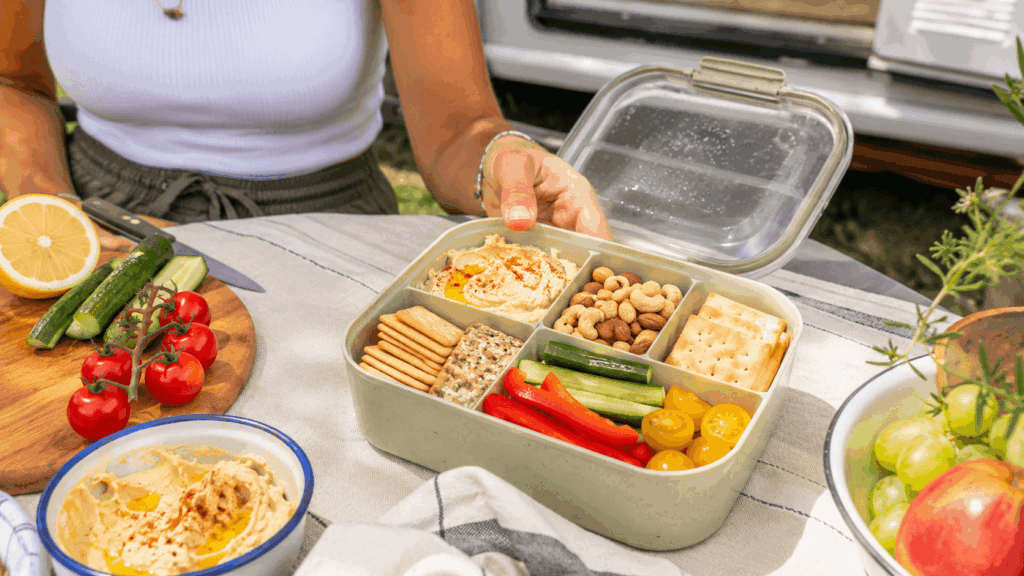 Geöffnete Snackbox mit Hummus, Nüssen, Gemüse und Crackern auf einem Campingtisch vor einem Wohnwagen