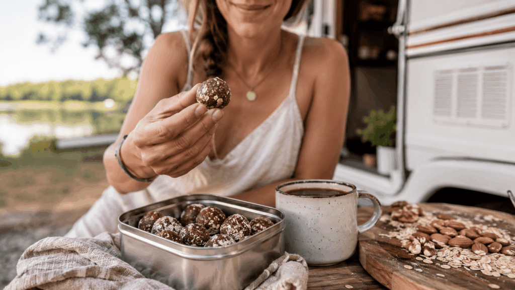 Eine Frau hält einen Kakao-Bliss-Ball vor einer Dose mit Bliss Balls, Nüssen und Haferflocken beim Camping.