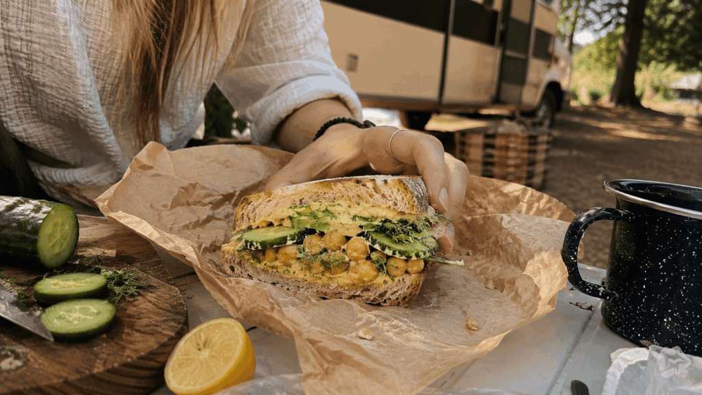 Ein Kichererbsen-Sandwich mit Gurke und Kräutern liegt auf Backpapier in einer Campingumgebung.